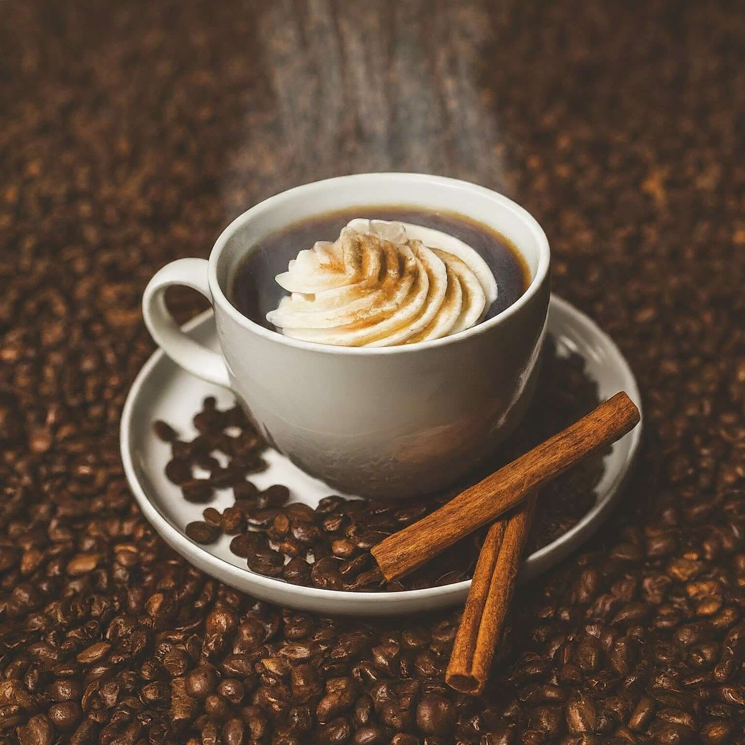 Cup of coffee with whipped cream surrounded by coffee beans and cinnamon sticks